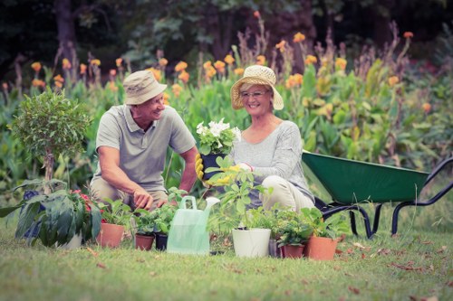 A mature man and woman sit together in a well-maintained garden, surrounded by gardening pots and tools, both wearing light-colored hats and casual clothes. They are engaging with various potted plants, including flowering and foliage varieties, positioned on the grass in front of them. Behind them, a lush background features a mix of flowering plants with purple, orange, and yellow blooms, as well as green foliage, creating a vibrant and colorful outdoor setting. To one side, a black garden bench is partially visible, and a green garden watering can is placed among the pots on the grass. The environment appears to be an outdoor residential garden in sunlight, suggesting a peaceful, well-cared-for space designed for gardening activities, with natural tones of soil, leaves, and blooming flowers illustrating a thriving, lively garden scene in Soho or nearby in London.