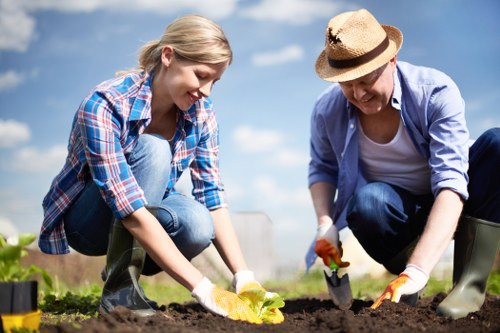A young woman and an older woman are working together in a garden under a bright blue sky with scattered clouds. They are kneeling on the soil, planting a small yellow-leaved plant, each holding gardening tools. The young woman is wearing a blue and red plaid shirt over a light top with blue jeans and grey gardening boots. The older woman is dressed in a light blue jacket, white top, and dark blue jeans, completed with a straw hat and orange gardening gloves. The garden features well-maintained, dark, enriched soil with a neat border of green plants and grass nearby, indicating a landscaped outdoor space. In the background, there are a few neatly trimmed bushes and trees, suggesting a residential or community garden setting in Soho or a similar area. The scene is naturally lit, capturing a pleasant and productive gardening moment, consistent with landscaping and lawn care activities carried out by professionals like Landscapers Soho.