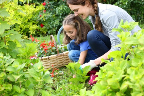 A woman and a young girl are gardening together in a lush, well-maintained outdoor space, with vibrant green foliage and a variety of flowering plants surrounding them. The woman is kneeling on the grass, leaning forward to assist the girl as she gathers red flowers into a woven wicker basket. The garden features a neatly edged flower bed with rich, dark soil and dense greenery, including broad-leaved shrubs and small trees in the background, creating a natural and inviting environment. Sunlight filters through the leaves, illuminating the scene with warm, natural light, suggesting a mild, clear day. The scene reflects the care and attention involved in outdoor gardening and landscaping, highlighting the services offered by landscapers in Soho, which include flower bed creation and garden maintenance, supporting local expertise in creating attractive and sustainable outdoor spaces.