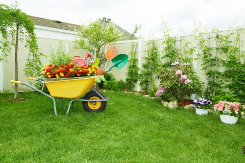 A well-maintained residential garden with a lush, green lawn occupying the foreground, edged by flower beds and potted plants full of blooming pink, purple, and white flowers. In the middle of the garden, a bright yellow wheelbarrow filled with colourful garden decor, including a pink flamingo ornament, is positioned on the grass, alongside garden tools such as a green watering can and a blue spade. The background features a white wooden fence with climbing green ivy and foliage, with a few trees and shrubs providing natural height and structure. The sky above is partly cloudy, casting soft, natural light across the outdoor space, which appears neat and inviting, ideal for outdoor gardening activities or relaxation. This scene illustrates typical gardening and landscape maintenance elements common to properties serviced by landscapers in Soho, suitable for showcasing professional garden care in the local area.
