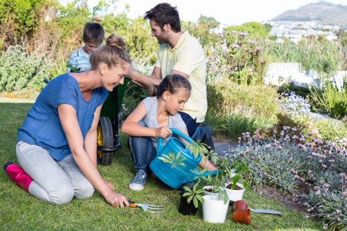 A family of four, including two children and two adults, engage in gardening activities in a well-maintained backyard garden. The garden features a lush, green lawn with neatly trimmed grass, bordered by flower beds filled with colorful blooms and various plants. In the foreground, the woman is kneeling on the grass, planting or tending to a small shrub, with gardening tools and a small white watering can nearby. The girl is sitting on the lawn, holding a blue backpack filled with garden tools and watering cans, actively participating in gardening tasks. The father and son are standing behind, with the father observing and the boy holding a small gardening tool, both focused on the gardening activity. The garden includes a paved area or stepping stones leading through the space, with dense shrubbery, flowering plants, and possibly a tree in the background. The scene is illuminated by natural daylight under clear weather, creating a bright and inviting outdoor environment typical of a residential garden in Soho or nearby areas, demonstrating outdoor maintenance and family gardening efforts by landscapers Soho.