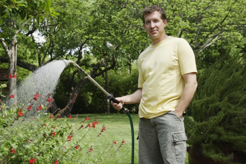 A man with light brown hair, wearing a pale yellow T-shirt and grey cargo shorts, stands in a well-maintained garden during daytime, holding a garden hose and watering the plants and flower beds in front of him. The garden features a lush, green lawn with dense shrubbery and trees in the background, including a neatly trimmed hedge on the right side. The flower beds contain small red blossoms, and the area appears tidy and cared for, illustrating typical outdoor gardening activities that a professional landscaper from Landscapers Soho might undertake to enhance garden aesthetics and health. The weather looks mild and overcast, with soft natural lighting illuminating the scene, emphasizing the outdoor environment’s vibrant greenery and flourishing plant life consistent with a typical garden in the Soho area near central London, suggesting local expertise in gardening and outdoor care.