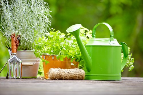 A green watering can is placed on a wooden outdoor table alongside potted herbs, including a lavender plant in a beige pot and a bright green leafy plant in a terracotta pot. The scene also features a coiled garden twine and a small gardening hand tool with a wooden handle and metal forked head, all set against a blurred background of lush green foliage, suggesting a well-maintained garden space typical of a residential outdoor area in Soho. The natural lighting highlights the vibrant green tones of the plants and garden tools, emphasizing outdoor gardening activities supported by Landscapers Soho's planting and garden maintenance services in the local area, with a focus on cultivating attractive, healthy garden spaces.