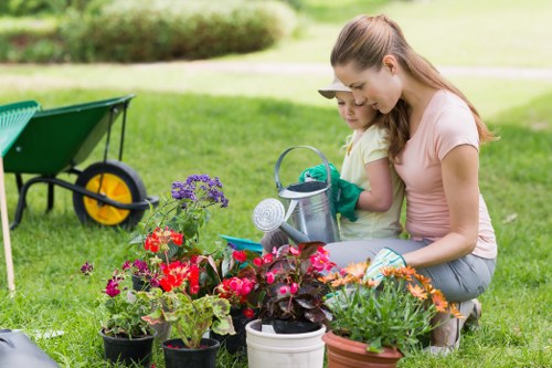 In a well-maintained garden with lush green grass typical of Soho areas, a young woman and a girl are engaged in planting and caring for a variety of flowering plants and foliage in an outdoor space. The garden features a neatly trimmed hedge in the background and a paved or gravel pathway partially visible nearby. The foreground is filled with vibrant potted plants, including mixed flowers with red, purple, and orange blooms, arranged around the women. A wheelbarrow with a green body and yellow wheel sits on the grass on the left side, indicating ongoing gardening work. The woman is wearing a light pink t-shirt and jeans, while the girl is dressed in a yellow top, both focused on the plants, with the woman guiding or assisting the child. The scene reflects a calm, productive moment in a typical residential garden, emphasizing natural tones, plant variety, and garden maintenance activity, reminiscent of gardening services provided by local landscapers in Soho.