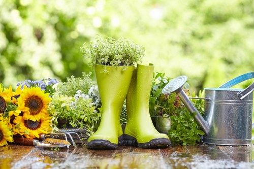 A garden scene featuring a wooden outdoor table with various gardening items arranged on it, including a pair of bright green rubber gardening boots filled with small white and green flowering plants, alongside a small bouquet of sunflowers and other assorted flowers with yellow, purple, and green foliage. To the right, there is a silver metal watering can with a long spout and a handle, set against a blurred background of lush green trees and foliage, indicating an outdoor, well-maintained garden space typical of domestic gardens in Soho area. The overall lighting suggests a bright, sunny day, illuminating the natural colours and textures of the plants, tools, and wooden surface. This scene illustrates typical gardening preparations or outdoor plant care in a landscaped backyard or front garden.