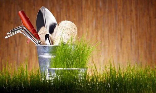 The image features a close-up view of a galvanized metal bucket and a smaller container placed on a well-maintained grassy lawn, with several gardening tools arranged inside the larger bucket, including a small hand rake with a wooden handle, a trowel, and a garden fork. There is a spool of natural twine or string and a clump of vibrant green grass or young plants positioned within the containers. The background is a wooden surface with warm, natural tones, providing a simple, rustic setting that highlights the gardening tools and plant elements. The scene emphasizes outdoor gardening activities, suitable for gardening or landscaping services in the Soho area, and showcases a neat, organized approach to outdoor plant care and lawn maintenance, aligned with professional gardening standards. Natural lighting suggests an overcast day or shaded outdoor environment, enhancing the detailed textures of the tools, grass, and wooden background.