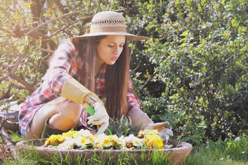 A young woman wearing a wide-brimmed straw hat, plaid shirt, and gardening gloves is kneeling on the grass in a well-maintained outdoor garden space. She is carefully tending to a flower bed filled with vibrant yellow and purple flowers, which are bordered by a circular edging. The garden features lush green grass, dense hedge lines, and leafy trees in the background, creating a natural and serene environment typical of gardens in Soho. Natural sunlight illuminates the scene, highlighting the textures of the plants, soil, and the wooden garden edging. This outdoor setting exemplifies careful garden maintenance, with a focus on planting and nurturing flowers, aligning with the gardening services offered by Landscapers Soho, especially within the context of sustainable gardening practices outlined on their recycling and sustainability page.