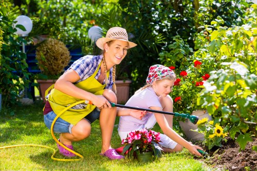 A woman and a young girl are gardening together in a lush, well-maintained backyard garden in Soho. The woman, wearing a straw hat, yellow apron, and casual clothing, is crouched down smiling as she helps the girl tend to a flower bed. The girl, dressed in a floral hat and light clothing, is focused on planting or watering flowers, with a gardening hose connected to her hand. The garden features a neatly mown lawn, vibrant flower borders with colorful blooms, including red, yellow, and pink flowers, and lush green foliage with some trees and shrubs in the background. Decorative garden lights are visible hanging among the plants, and the overall environment appears sunny and inviting, ideal for outdoor gardening and landscaping activities. This scene highlights active gardening efforts that could be supported by professional landscaping and garden maintenance services, such as those offered by Landscapers Soho, in the Soho area or nearby London postcode.